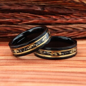 May include: Two black rings with gold accents and a textured design. The rings have a gold band in the center with gold flakes. The rings are set on a wooden surface.