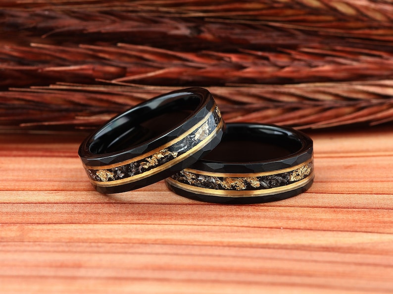 May include: Two black rings with gold accents and a textured design. The rings have a gold band in the center with gold flakes. The rings are set on a wooden surface.