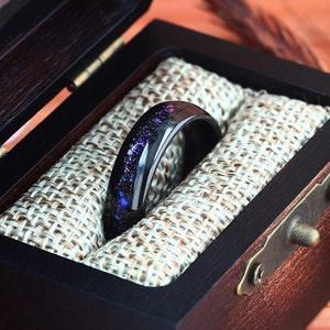 Pu&ograve; includere: Un anello nero con un design a galassia viola e blu all'interno di una scatola per anelli in legno con una fodera in tessuto.