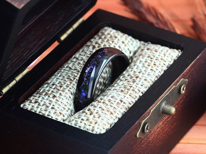 Pu&ograve; includere: Un anello nero con un design a galassia viola e blu all'interno di una scatola per anelli in legno con una fodera in tessuto.