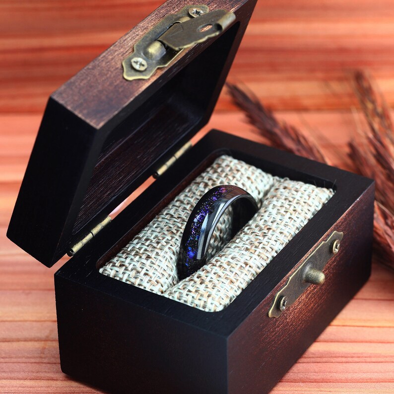 Pu&ograve; includere: Un anello nero con glitter viola e argento incorporati nella resina &egrave; esposto in una scatola per anelli in legno con un rivestimento in velluto marrone.
