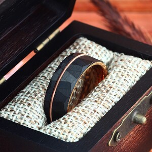 Puede incluir: Un anillo de carburo de tungsteno negro con una incrustaci&oacute;n de oro rosa y un acabado martillado, presentado en una caja de madera con un forro de tela.