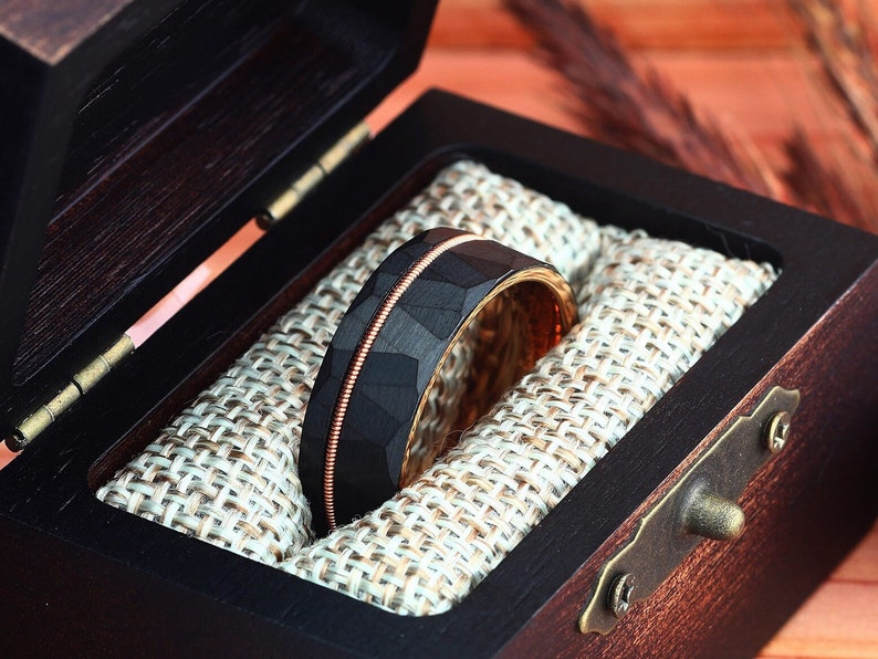 Puede incluir: Un anillo de carburo de tungsteno negro con una incrustaci&oacute;n de oro rosa y un acabado martillado, presentado en una caja de madera con un forro de tela.