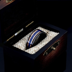 May include: A black tungsten ring with blue and brown wood inlay. The ring is in a wooden box with a brown fabric lining.