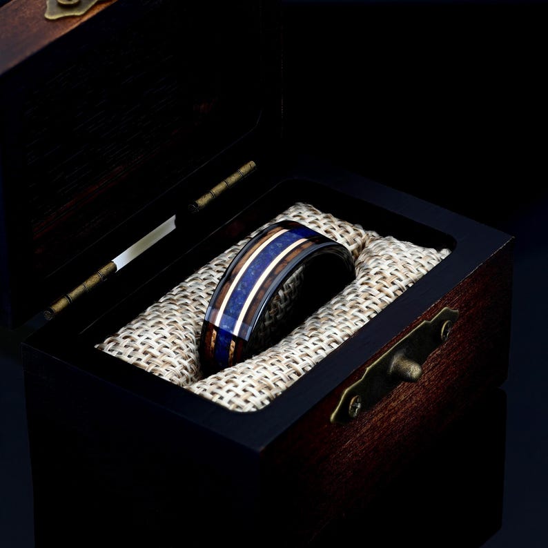 May include: A black tungsten ring with blue and brown wood inlay. The ring is in a wooden box with a brown fabric lining.