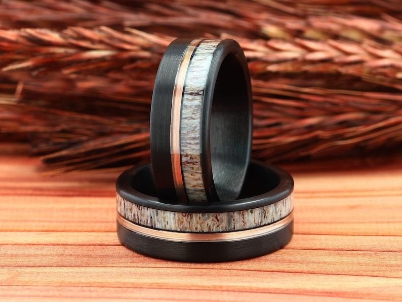 Puede incluir: Dos anillos de fibra de carbono negra con una banda fina de oro rosa y una incrustaci&oacute;n de asta blanca. Los anillos est&aacute;n apilados uno encima del otro.