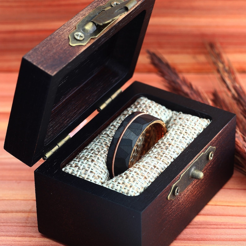 Puede incluir: Un anillo de carburo de tungsteno negro y oro rosa con acabado martillado, presentado en una caja de anillo de madera con forro de terciopelo marr&oacute;n.