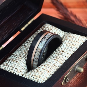 Puede incluir: Un anillo negro y marr&oacute;n con una banda de cobre y una incrustaci&oacute;n de madera en una caja de anillo de madera con un forro de arpillera.