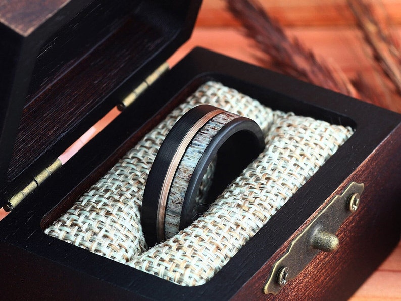 Puede incluir: Un anillo negro y marr&oacute;n con una banda de cobre y una incrustaci&oacute;n de madera en una caja de anillo de madera con un forro de arpillera.