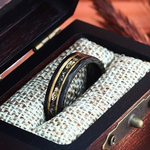 May include: A black ring with gold and dark accents, presented in a dark wooden box with a light-colored fabric lining. The ring features a band of gold and black materials, with a textured design. The box is open, revealing the ring's intricate details.