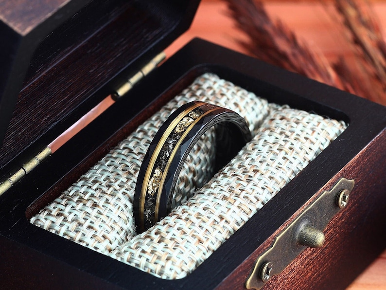 May include: A black ring with gold and dark accents, presented in a dark wooden box with a light-colored fabric lining. The ring features a band of gold and black materials, with a textured design. The box is open, revealing the ring's intricate details.