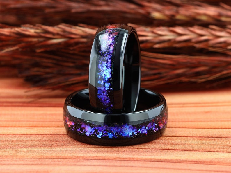 Pu&ograve; includere: Due anelli in ceramica nera con un'incisione di glitter viola e blu. Gli anelli sono impilati uno sopra l'altro.