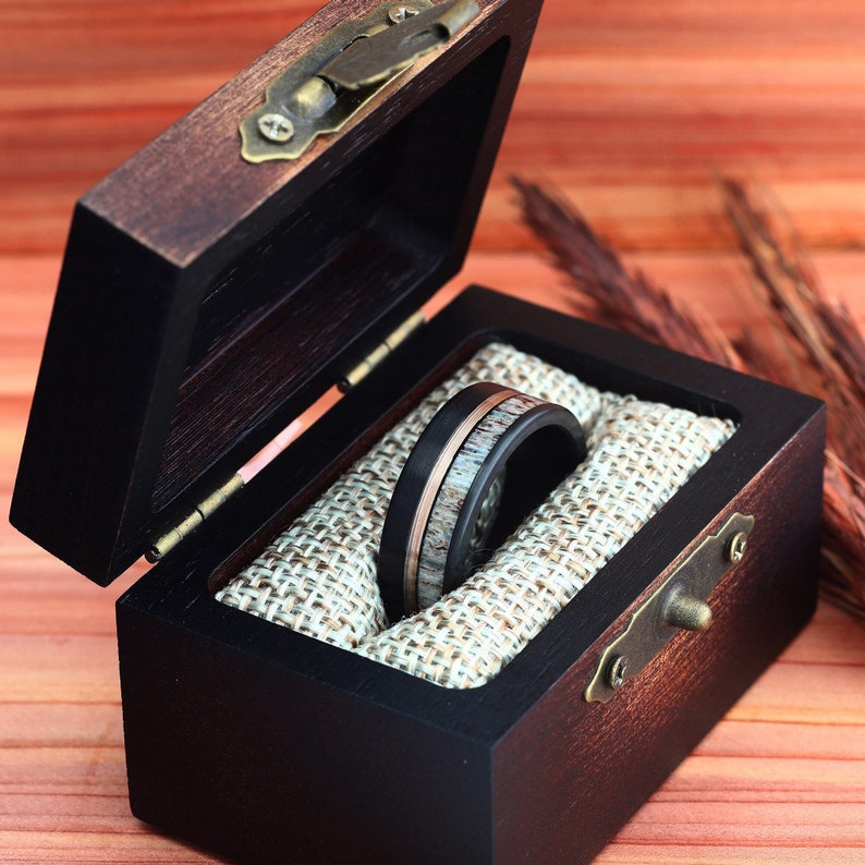 Puede incluir: Un anillo negro y marr&oacute;n con una banda de cobre se encuentra en una caja de anillo de madera con un forro de terciopelo marr&oacute;n.