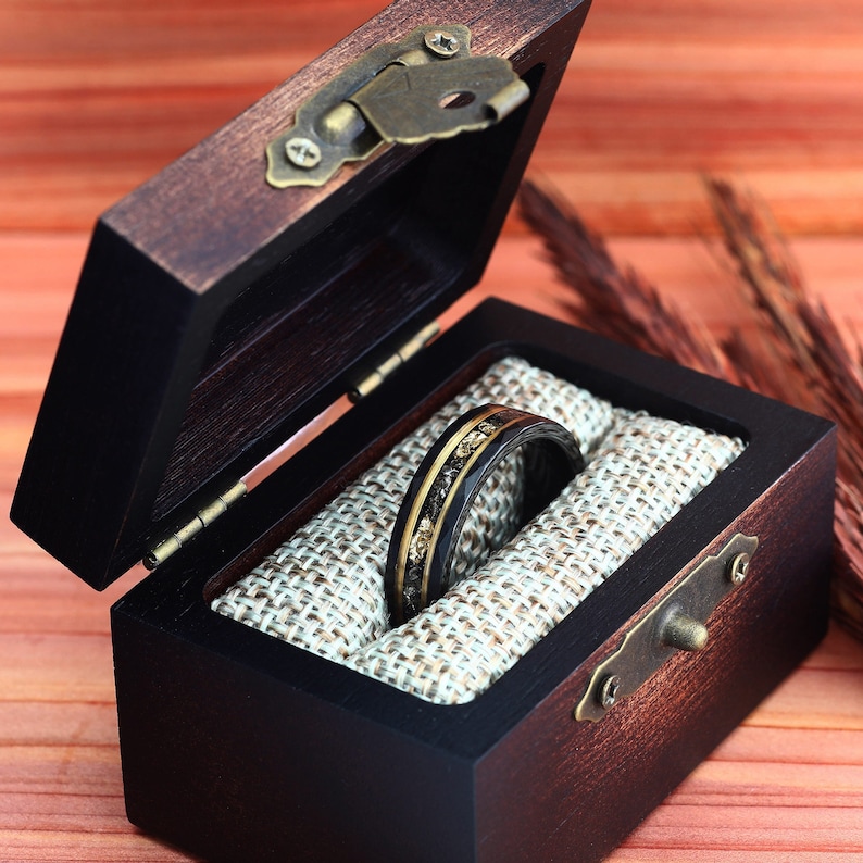 May include: A dark wooden ring box with a hinged lid, open to reveal a black ring with gold and silver accents. The ring rests on a beige fabric lining. The box has a brass clasp and hinges.