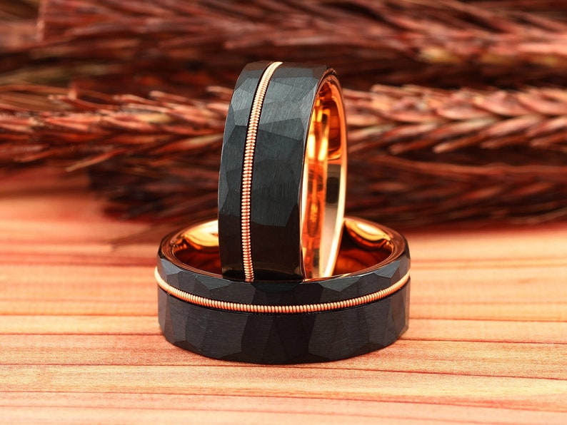 Puede incluir: Dos anillos de carburo de tungsteno negro con una incrustaci&oacute;n de oro rosa. Los anillos tienen un acabado martillado y est&aacute;n apilados uno encima del otro.