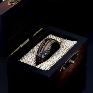 May include: A black tungsten ring with a brown wood inlay and a silver band. The ring is in a wooden box with a brown fabric lining.