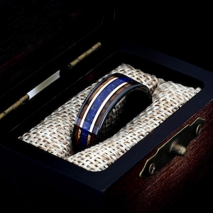 May include: A black tungsten ring with a blue and brown wood inlay. The ring is in a wooden box with a brown fabric lining.