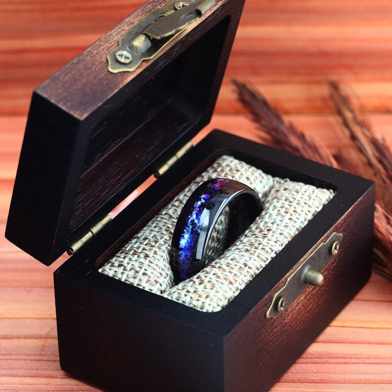 Pu&ograve; includere: Un anello nero con un design a galassia blu e viola &egrave; esposto in una scatola per anelli in legno con un rivestimento in velluto marrone.