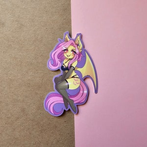 May include: A sticker featuring a cartoon character with pink hair and yellow fur, dressed in a black lingerie outfit with bat wings. The character has a mischievous expression and is sitting with one leg bent.