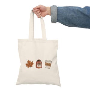May include: A cream-coloured canvas tote bag featuring a printed graphic of a brown maple leaf, a small orange candle labelled Pumpkin Spice, and a white disposable coffee cup with a brown sleeve, arranged horizontally across the centre.