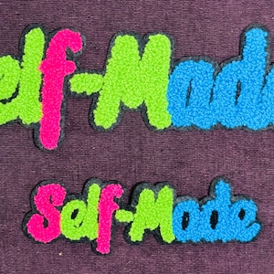 Puede incluir: Dos parches bordados con las palabras "Self-Made". El parche superior, más grande, presenta letras rosas, verdes y azules. El parche inferior, más pequeño, repite la misma combinación de colores. Ambos tienen un borde negro sobre un fondo morado.