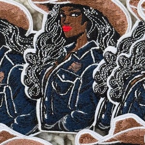 Könnte beinhalten: Gestickter Aufnäher mit einer dunkelhäutigen Frau, die einen braunen Cowboyhut und eine dunkelblaue Jeansjacke trägt. Ihr langes, welliges schwarzes Haar umrahmt ihr Gesicht, und sie hat roten Lippenstift. Der Aufnäher hat einen weißen Rand.