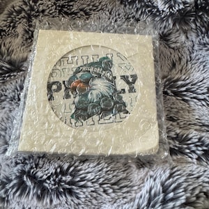 May include: A square, off-white item featuring a circular design with the text "PHILADELPHIA" and "EAGLES" in a green and white color scheme. The item is wrapped in bubble wrap and rests on a gray and white textured surface.
