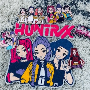 Puede incluir: Una colección de parches bordados coloridos con ilustraciones de personajes femeninos de estilo caricatura. Los parches incluyen la palabra "HUNT RX" y el nombre "MIRA". Los parches están sobre una superficie texturizada de color gris claro.