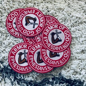 May include: A collection of red, circular embroidered patches. Each patch features the text "ARMOR OF GOD EPHESIANS 6:11" in white, surrounding a central design of a brown arm holding a cross. The patches are scattered on a textured, off-white surface.