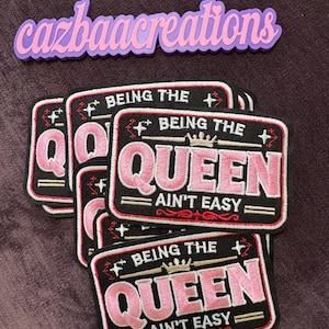 May include: A stack of rectangular embroidered patches with a black background and pink lettering that reads Being The Queen Ain't Easy. Each patch features a gold crown icon, white stars, and red decorative flourishes. The text cazbaacreations is visible.