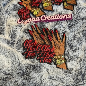 Może przedstawiać: Haftowane naszywki przedstawiające dłoń z czerwonymi paznokciami, złotym zegarkiem i napisem "Clock That Tea". Naszywki są brązowe, czerwone i czarne, ze złotymi akcentami. Naszywki znajdują się na szarej i białej puszystej powierzchni. Widoczny jest również napis "Cypaa Creations".