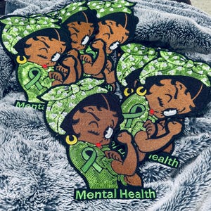 Könnte beinhalten: Aufnäher mit einer Cartoonfigur mit grünem und weißem Pailletten-Kopftuch und grünem Band. Die Aufnäher tragen die Worte "Mental Health". Die Aufnäher befinden sich auf einer grauen, flauschigen Oberfläche.