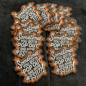 May include: A pile of black embroidered patches. Each patch features the phrase "Sometimes You Gotta Pop Out And Show #$$!!??!" in white and orange, with an orange sunburst design.
