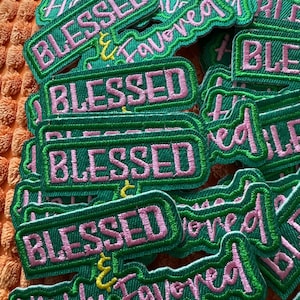 Pode incluir: Uma coleção de patches bordados verdes com letras rosa. Os patches apresentam as palavras "BLESSED" e "Highly Favored" em uma fonte cursiva. Os patches têm uma borda verde e estão espalhados em uma superfície laranja texturizada.