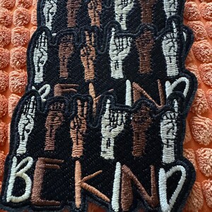 May include: Black embroidered patches with various hand gestures in white and brown, with the words "BE KIND" in white and brown thread. The patches are stacked on an orange textured surface.