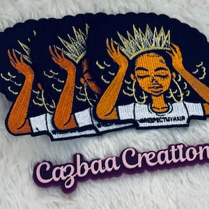 May include: Embroidered patch featuring a Black woman with a gold crown, orange arms, and black hair. The woman is wearing a white shirt with the text "RESPECTMYHAIR". The patch is black with a scalloped edge. A pink and purple "Cazbaa Creations" logo is below.