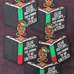 以下が含まれることがあります： 黒人女性がターバンを巻いて開いた本を持っている刺繍パッチ。本には「OUR HISTORY... TOO STRONG TO GO UNTOLD!」というテキストが白で書かれており、緑と赤のアクセントがあります。パッチは濃い紫色の背景にあります。