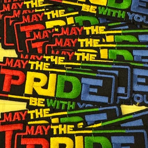 May include: Embroidered patches with the text "MAY THE PRIDE BE WITH YOU." The patches feature the words in rainbow colours: red, yellow, green, and blue, on a black background. The patches are rectangular.
