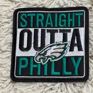 May include: A square embroidered patch with a black border featuring the text STRAIGHT in teal, OUTTA in black, and PHILLY in teal. A white, black, and teal eagle head logo is positioned in the center over the word OUTTA on a white background.
