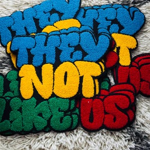 以下が含まれることがあります： 「THEY NOT LIKE US」という文字が太いバブルスタイルのフォントでデザインされた、タフティング生地のワッペンコレクション。ワッペンは重ねられており、青、黄、緑、赤の鮮やかな色合いで、それぞれ太い黒の縁取りが施されています。