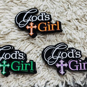 May include: Three embroidered patches featuring the text God's Girl in white and colored thread. Each patch includes a cross icon with a small white dot pattern. One patch has an orange cross and text, one has green, and one has light purple.