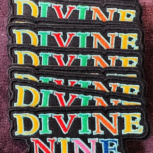 May include: A stack of black embroidered patches with the words "DIVINE NINE" in colorful, bold lettering. The letters are outlined in a light blue, with a variety of colors filling each letter, including yellow, green, orange, and pink.