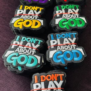Pode incluir: Patches bordados com o texto "I DON'T PLAY ABOUT GOD" em várias cores, incluindo roxo, vermelho, amarelo, verde, turquesa, azul e laranja. Os patches são pretos com detalhes em branco e cinza e são presos com elásticos vermelhos.
