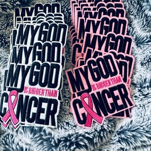 Pode incluir: Remendos bordados com o texto "MY GOD IS BIGGER THAN CANCER". Os remendos apresentam uma fita rosa e são em preto e branco ou rosa e preto. Os remendos são empilhados em uma superfície fofa, cinza e branca.