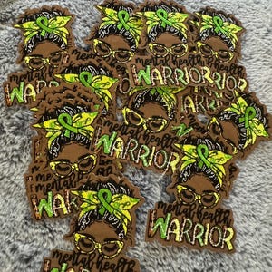 Può includere: Una collezione di toppe marroni raffiguranti una donna con occhiali e un nastro verde, con le parole "Mental Health Warrior". Le toppe hanno una combinazione di colori giallo e verde, e il testo è in un carattere stilizzato. Le toppe sono sparse su una superficie grigia.