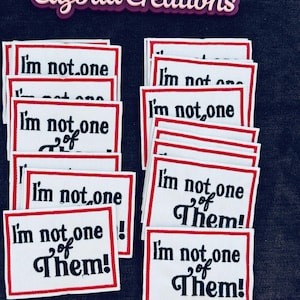 Peut inclure: Plusieurs écussons rectangulaires blancs avec des bordures rouges, affichant le texte "I'm not one of Them!" en noir. L'ensemble est présenté sur un fond sombre, avec le nom de la marque "Cazbaa Creations".