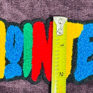 May include: A colorful embroidered patch with the word "ANIMATED" in bright yellow, red, blue, and green letters. The patch is approximately 2 inches long and has a black border. The patch is on a purple background.