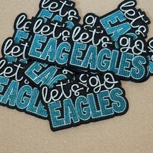 May include: A pile of embroidered patches with the phrase "Let's Go EAGLES" in white script and teal block letters on a black background. The patches are stacked on a tan surface.