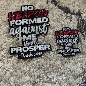 Puede incluir: Dos parches negros con el texto "NO WEAPON FORMED against ME shall PROSPER Isaiah 54:17". El texto "NO WEAPON" está en lentejuelas rojas, el resto en lentejuelas blancas. Un parche es más grande que el otro.