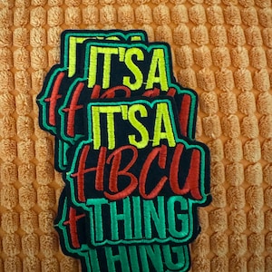 May include: Embroidered patch with the text "IT'S A HBCU THING" in green, red, and yellow on a black background. The patch is stacked on top of a textured orange fabric.